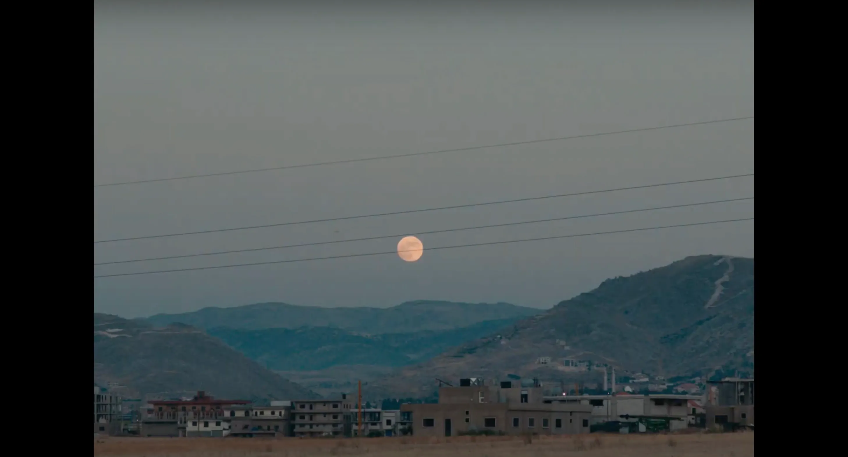 A pale moon rises over a hillside town and power lines at dusk